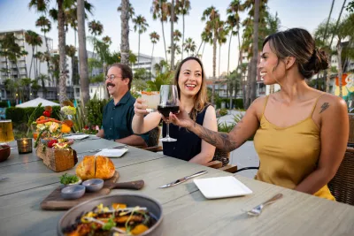 Three friends enjoying a meal outdoors, toasting with drinks amidst palm trees in San Diego, CA.