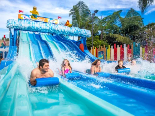 Six people race down parallel water slides on blue mats, creating splashes as they approach the end of the slides at Legoland in San Diego.