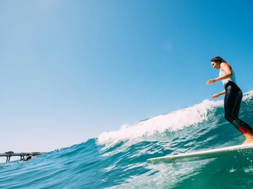 A person in a wetsuit rides a wave on a surfboard in la jolla, san diego near a pier under a clear blue sky.