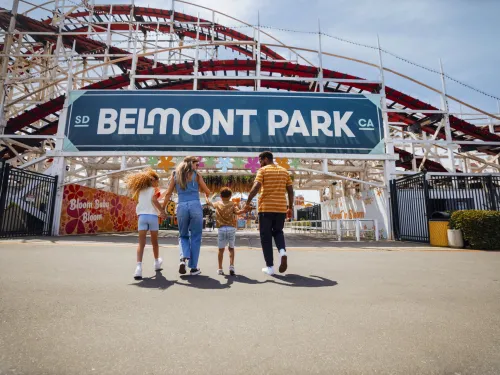 A family of four walks toward the entrance of Belmont Park in San Diego on Mission Beach, an amusement park with a large roller coaster structure visible behind the sign.