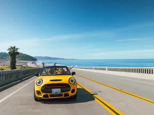 A yellow convertible with its top down drives on a coastal highway bridge next to the ocean in Torrey Pines under a clear blue sky, with passengers raising their arms.