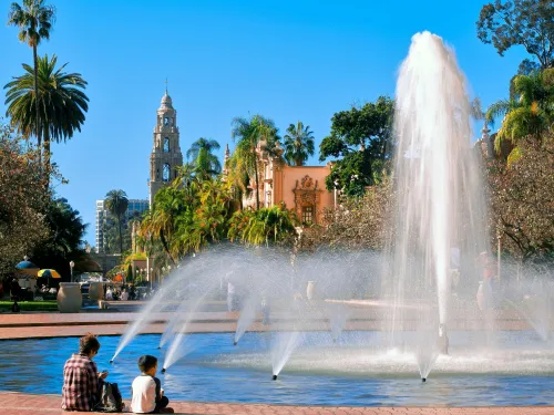 A large fountain sprays water in a park with palm trees, historic buildings, and clear blue sky; two people sit at the fountain's edge.