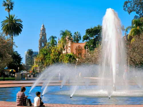 A large fountain sprays water in a park with palm trees, historic buildings, and clear blue sky; two people sit at the fountain's edge.