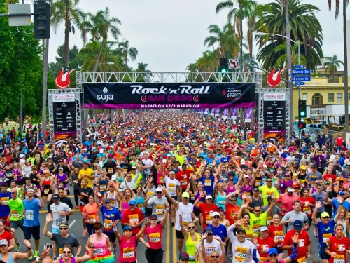 Large crowd of runners in colorful outfits starts the Rock 'n' Roll San Diego Marathon, with palm trees and banners visible in the background.