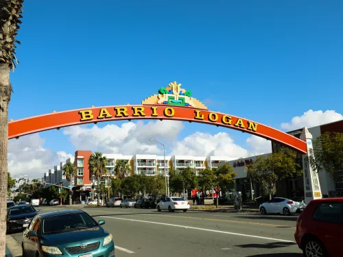 A large red arch with "Barrio Logan" written on it spans a street lined with parked cars and buildings under a blue sky.