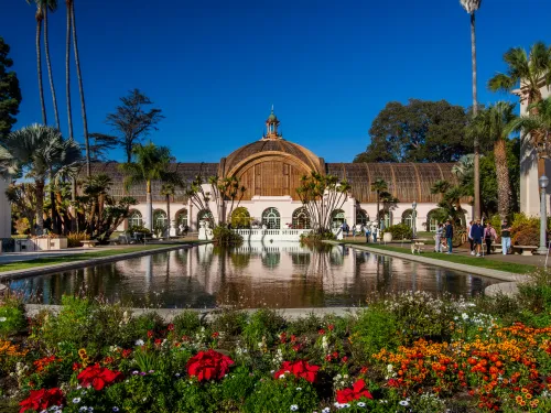 Balboa Park Botanical building with arched windows behind a reflective pond, surrounded by colorful flower beds, palm trees, and visitors on a clear day.