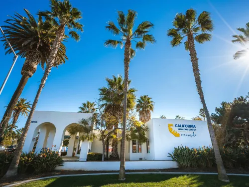 The California Welcome Center in Oceanside, a white building with arches, is surrounded by palm trees under a clear blue sky.
