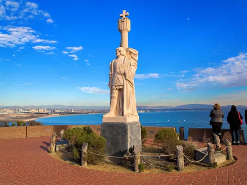 A stone statue stands on a raised platform overlooking the ocean, with four people observing the view and a cityscape visible in the background under a blue sky.