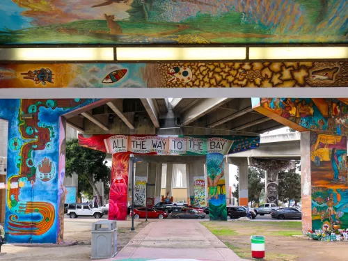 Colorful murals decorate the pillars and ceiling of an urban park area beneath a highway overpass, with benches, people, and trees visible in the scene.