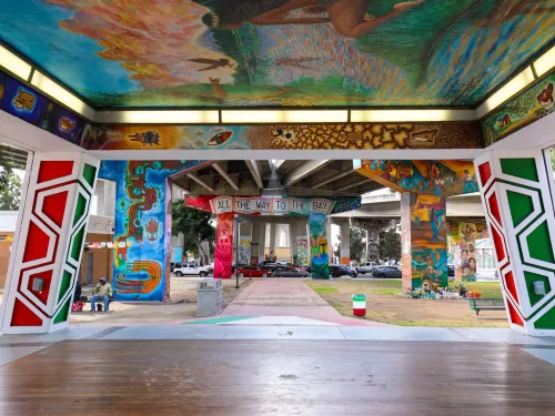 View from a stage under a mural-covered structure in a park, looking toward painted freeway pillars and a banner reading "All the way to the Bay.