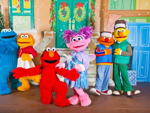 Six Sesame Street characters, including Cookie Monster, Elmo, Abby Cadabby, Ernie, and Bert, stand together in front of a festive backdrop with wreaths and a decorated Christmas tree.