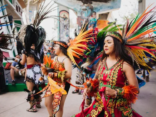 People in vibrant, traditional Aztec-inspired costumes perform a dance outdoors, surrounded by murals and a crowd of onlookers during Chicano Park Day.