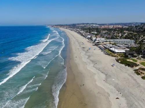 Aerial view of Del Mar's wide sandy beach with waves along the shoreline, people scattered on the sand, and buildings lining the coast under a clear blue sky.