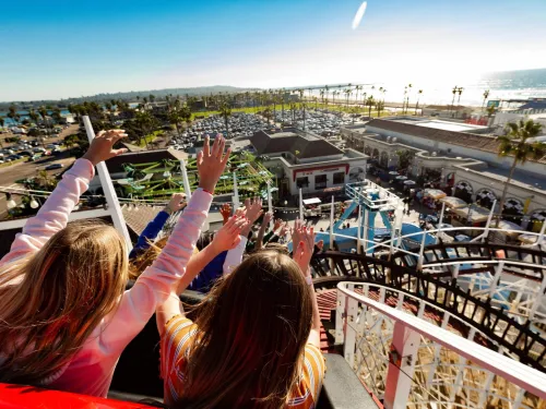 Three people with raised arms ride a wooden roller coaster at Belmont Park in San Diego on Mission Beach with a view of a parking lot, palm trees, and the ocean in the background on a sunny day.