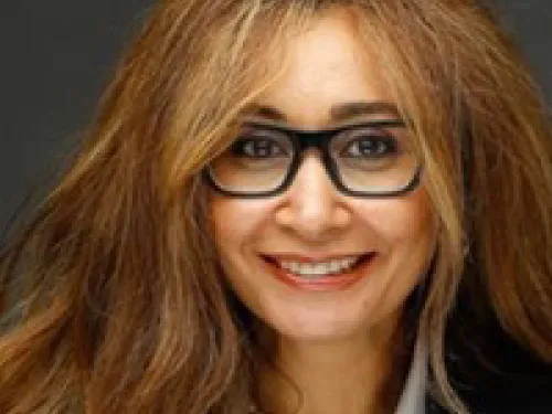 Hanaa Zahran with long wavy brown hair, wearing black glasses. a gray blouse, black blazer, and a pearl necklace, smiling at the camera against a dark background.