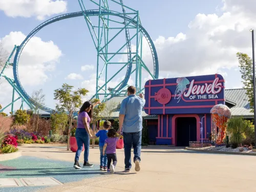 A family of four walks toward a shop called "Jewels of the Sea" near a large roller coaster on a sunny day at SeaWorld San Diego.