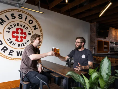 Two brewery patrons cheersing with the Mission Brewery Logo painted on the wall