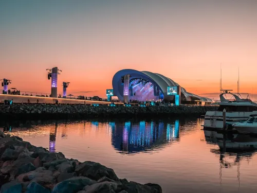 An outdoor concert venue with a modern, curved roof is lit up at sunset, reflecting in calm water with yachts docked nearby in San Diego.