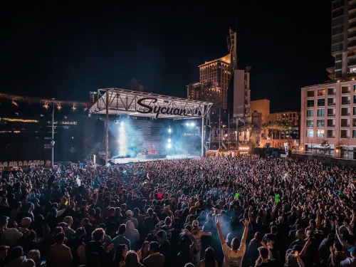 Large nighttime outdoor concert at Petco Park with a packed audience facing a lit stage labeled "SycamoR." Tall buildings surround the venue under a clear, dark sky.