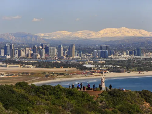 A group of people stands on a hill overlooking the San Diego skyline, with a beach, bay, and distant snow-capped mountains in the background.