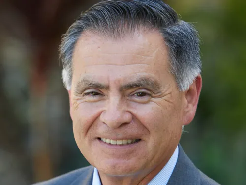 Rick Meza, smiling, wearing a striped tie, light blue undershirt and blue suit.