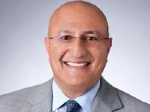 Sam Attisha wearing glasses, a light gray suit, blue shirt, and patterned tie poses in front of a plain gray background.