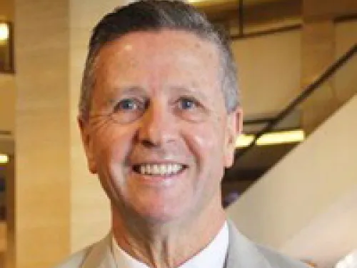Shaun Robinson in a light-colored suit and striped tie smiles at the camera in an indoor setting with blurred background lights.