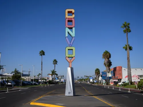 A tall, colorful neon sign reading "CONVOY" stands on a median in the middle of a wide street lined with palm trees and businesses under a clear blue sky.