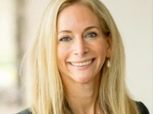 Lydia Bartell wearing a black blazer and blue top, smiles at the camera against a blurred light background.