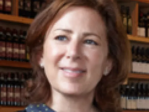 Tracy Borkum with shoulder-length brown hair stands in front of shelves stocked with bottles and wooden boxes, looking slightly to the side and smiling.