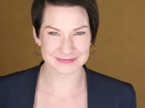 Annie Fitzgerald with short dark hair, wearing a navy blazer and white top, smiles at the camera against a plain brown background.