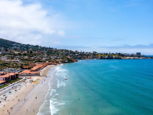 A coastal beach scene with blue water, sandy shore, people walking, and buildings along the shoreline under a partly cloudy sky.