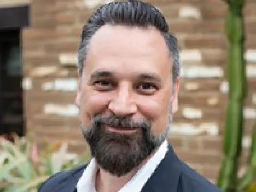 Eric Jenkins with dark hair and beard wearing a suit jacket and white shirt, smiling outdoors in front of a brick wall and plants.