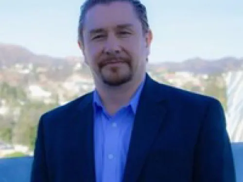Jamie Lemon with light skin, short brown hair, and a beard is wearing a dark suit and blue shirt, standing outdoors with a blurred cityscape and hills in the background.