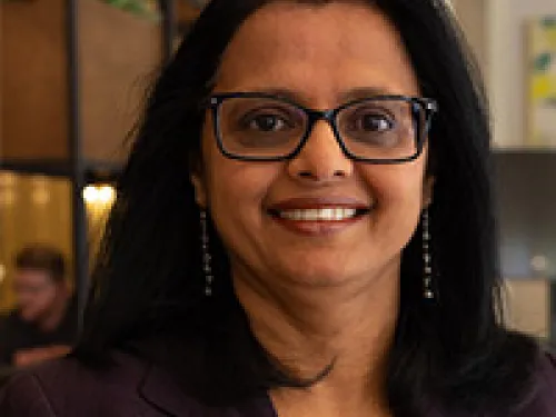 Cecilia Menezes with straight black hair, wearing glasses, a dark blazer, earrings, and a beaded necklace, smiling at the camera in an indoor office setting.