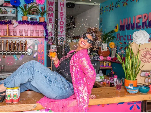 A woman in sunglasses poses on a bar counter, holding a drink and smiling, with confetti falling around her in a brightly decorated, colorful brewery.