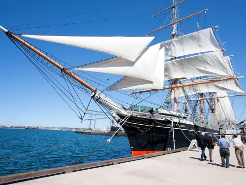 A large sailing ship with white sails docked at a pier, with four people standing nearby under a clear blue sky.