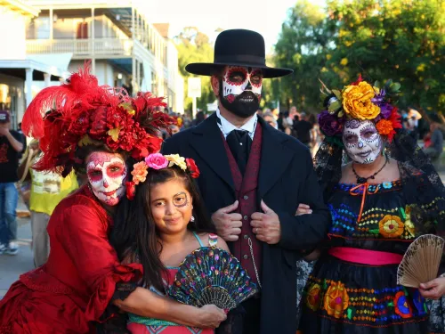 Four people dressed in colorful Day of the Dead costumes in San Diego and face paint stand together outdoors, with one young girl holding a decorative fan in front.