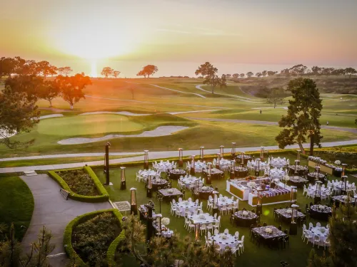 Outdoor event setup with round tables and chairs arranged on a lawn next to a golf course at sunset, with trees and a glowing sky in the background.