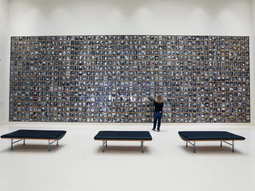 A person stands in front of a large wall display filled with many small framed photographs in a spacious, modern gallery with three black benches.