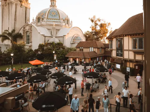 People gather in an outdoor courtyard with tables and umbrellas near historic buildings and a domed structure under a clear sky.