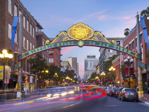The entrance arch to San Diego’s Gaslamp Quarter is shown over a busy street with blurred car lights and historic buildings at dusk.