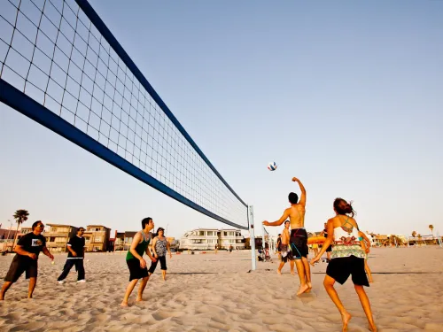 A group of people play beach volleyball on sand near the ocean, with houses and palm trees visible in the background under a clear sky.
