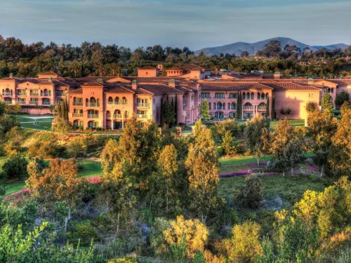 Large Mediterranean-style resort complex surrounded by lush trees and greenery, with distant hills in the background under a clear sky.