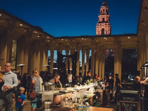 People gather at an outdoor bar and dining area in the evening; a tall illuminated tower is visible in the background against a dark blue sky.
