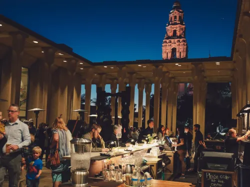 People gather at an outdoor bar and dining area in the evening; a tall illuminated tower is visible in the background against a dark blue sky.