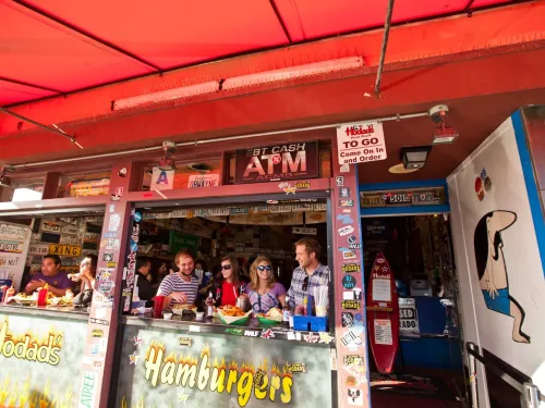 People sit at a counter eating food at an outdoor hamburger restaurant decorated with various signs and license plates under a red awning.