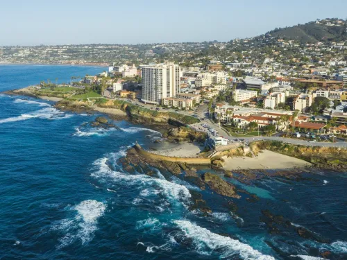 A coastal city with buildings along the shoreline, rocky cliffs, and waves crashing onto the beach under a clear blue sky.