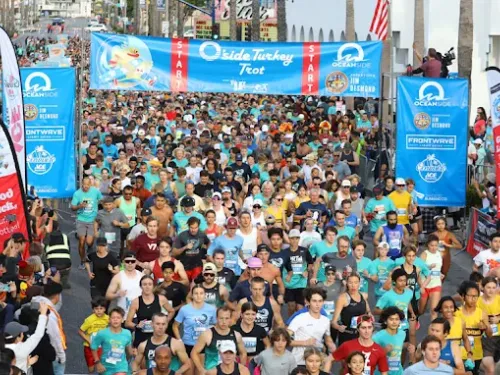 A large crowd of runners gathers at the starting line of the Surfside Turkey Trot race, with banners and sponsor signs visible in the background.