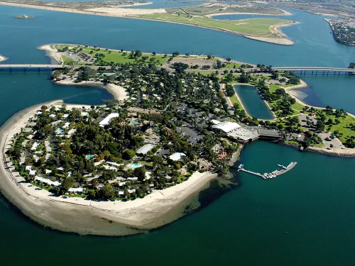 Aerial view of a small, lush island connected by a bridge, surrounded by water with sandy shores and greenery, featuring buildings and a dock.